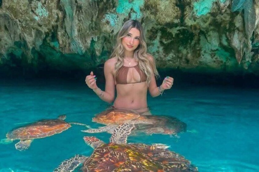 A person in a bikini surrounded by sea turtles in clear blue water with a rocky cave background. 5