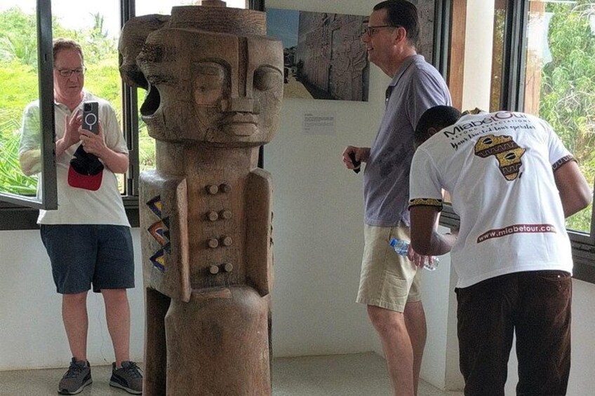 A large wooden statue with carvings, a person taking a photo, and another person in a white shirt with a logo. 2