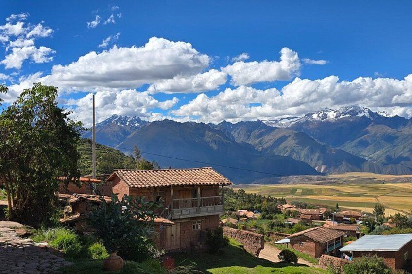 A rural village with terracotta-roofed houses, a mountain range with snow-capped peaks, and a clear blue sky with scattered clouds. 2