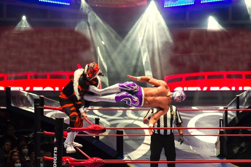 A wrestling match in a ring with a red and black theme, a referee, and an audience in the background. 3