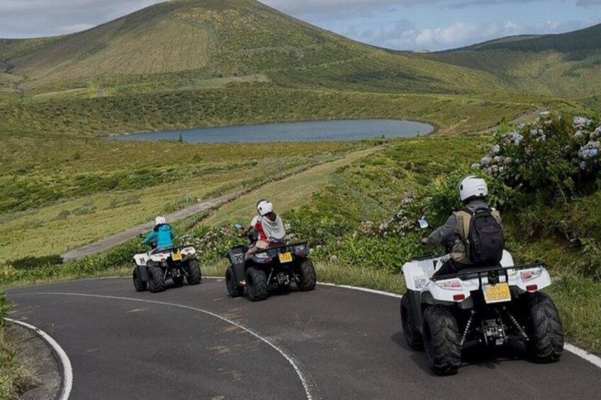 Three people riding ATVs on a winding road with a lake and hills in the background. 2