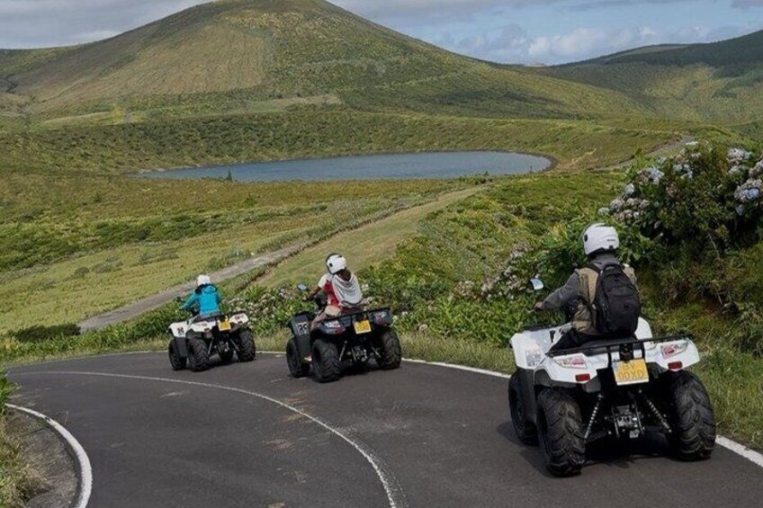 Three people riding ATVs on a winding road with a lake and hills in the background. 2