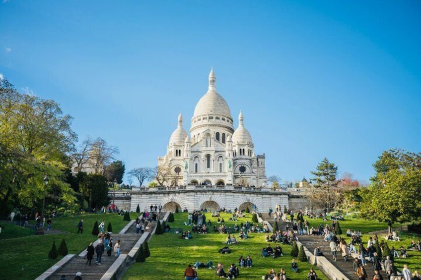 A large domed building with multiple smaller domes, surrounded by a park with people walking and sitting on the grass. 1