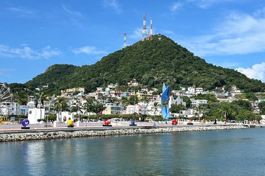A coastal cityscape with a hill, buildings, a pier, and a large blue sculpture. 1