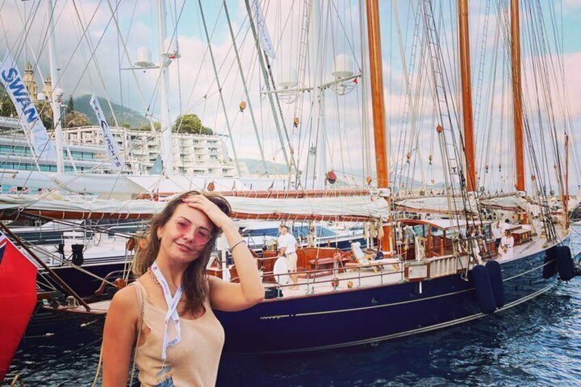 A person standing in front of a sailing ship with multiple masts and sails, docked at a marina with other boats and buildings in the background. 4