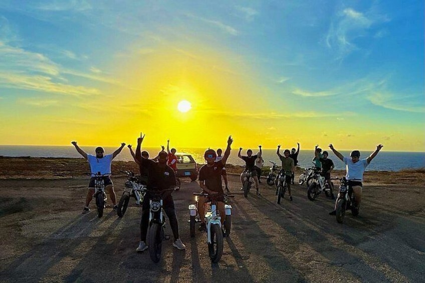 A group of people on bicycles on a dirt road with the sun setting in the background. 1