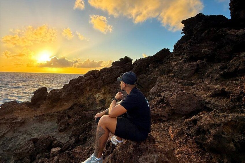 A person sitting on a rocky outcrop overlooking the ocean at sunset. 3