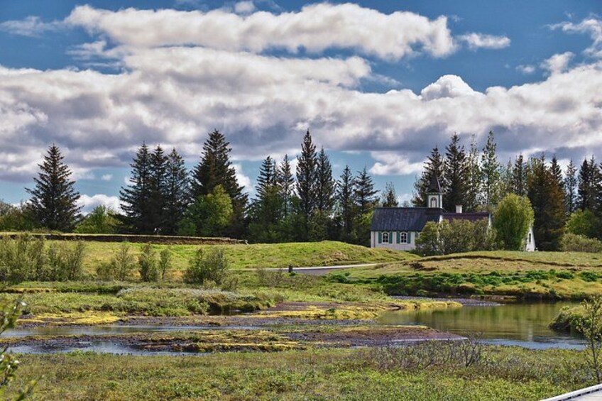 A rural landscape with a pond, a small white church, and a house surrounded by trees. 2