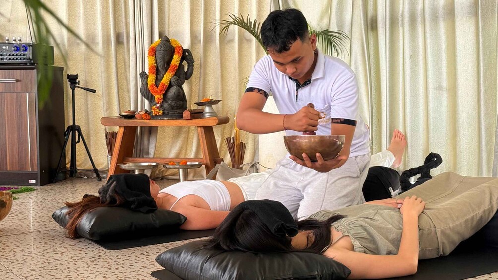 A person performing a ritual with a bowl and a statue, with two individuals lying on mats in a room with curtains and a tripod. 5