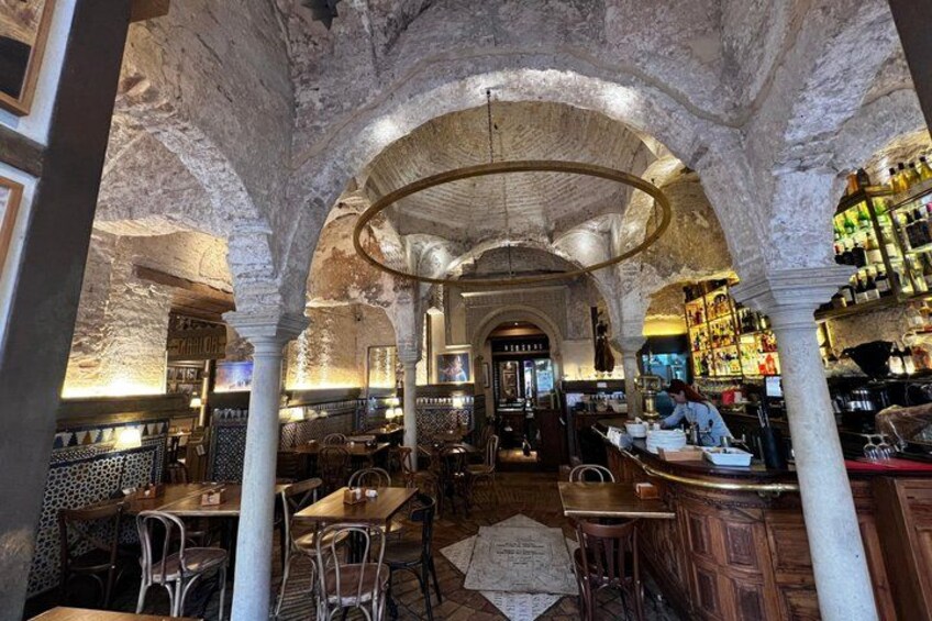 An interior with arched ceilings, wooden tables, and chairs, and a bar area with bottles. 5