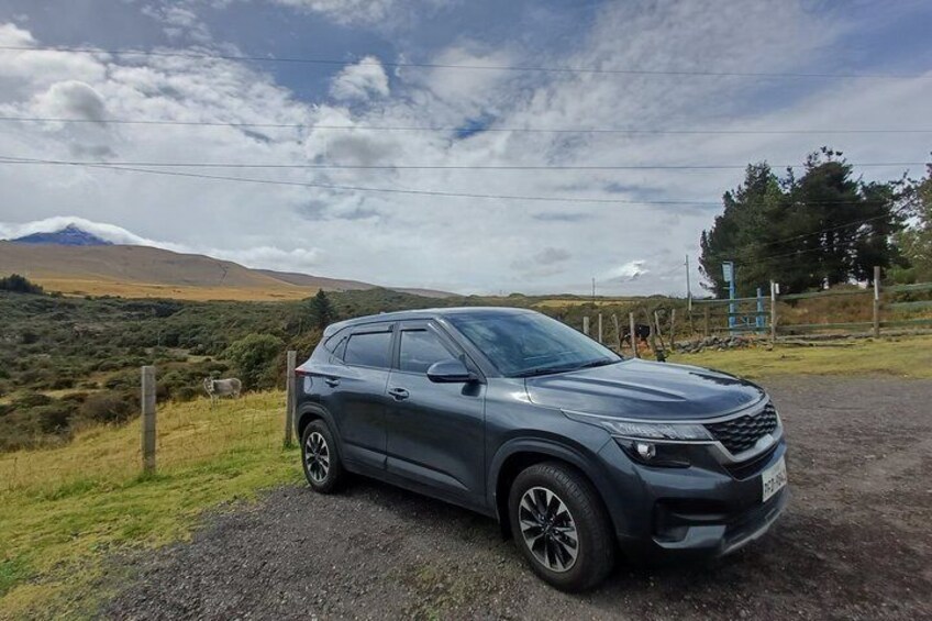 A dark-colored SUV parked on a gravel road with a rural landscape, including hills and a fence in the background. 4