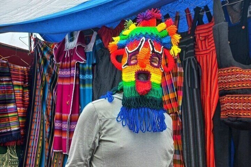 A person wearing a colorful, beaded mask with a fringe at the bottom, standing in front of a market stall with various colorful textiles. 1