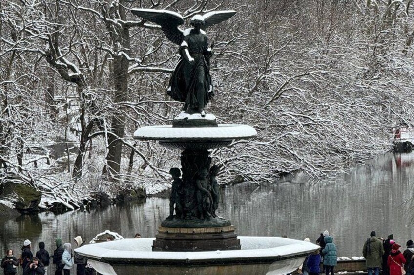 A winter scene with a statue of an angel on a fountain, surrounded by snow-covered trees and a calm body of water. 4