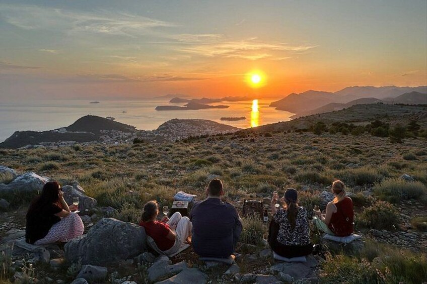 A group of people sitting on rocks at sunset overlooking a body of water and hills. 1