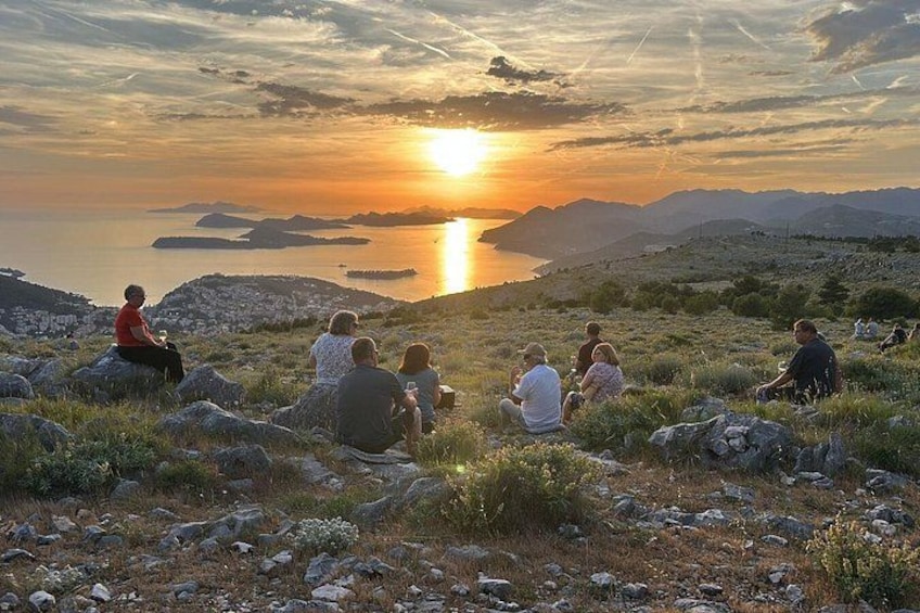 A group of people sitting on a rocky hillside overlooking a body of water during sunset. 4