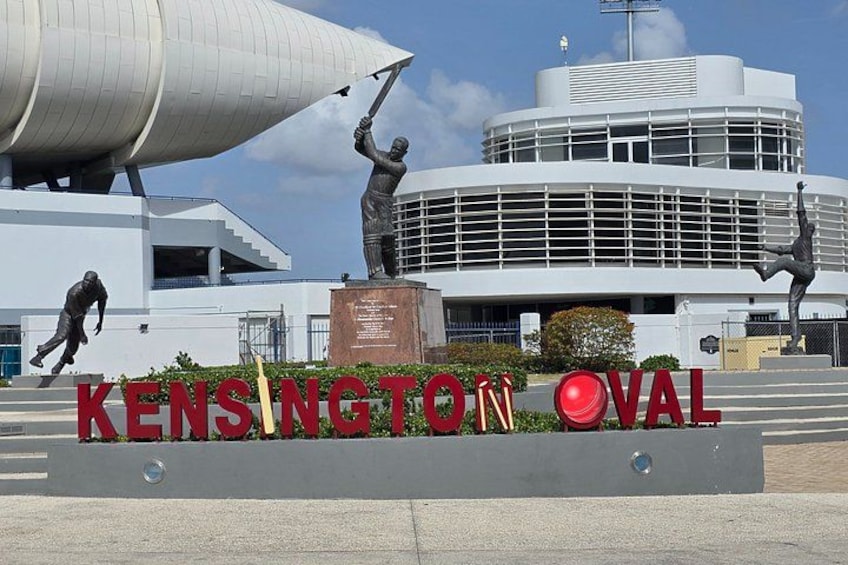A building with a distinctive circular design, a sign reading 'KENSINGTON OVAL,' and two statues of athletes. 1