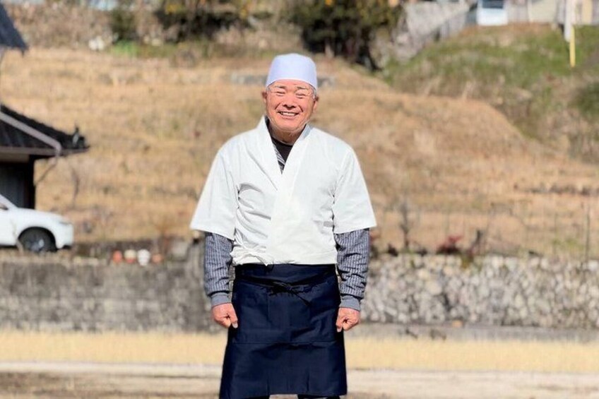 An elderly man in traditional attire standing outdoors with a rural background including a white car and a stone wall. 2