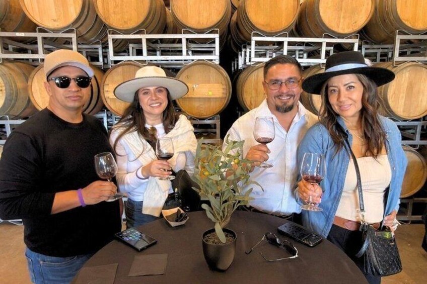 Four people standing at a table with wine glasses, surrounded by wooden barrels in a storage area. 2