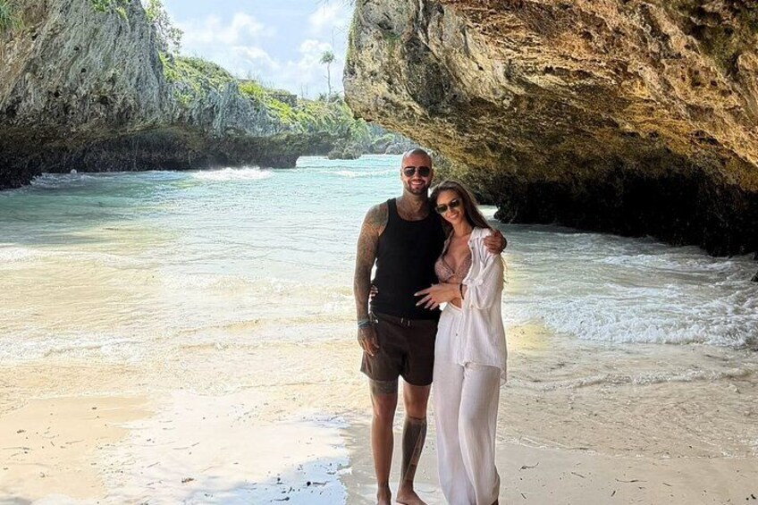 A beach with clear water, a rocky cliff, and a couple standing on the sand. 3