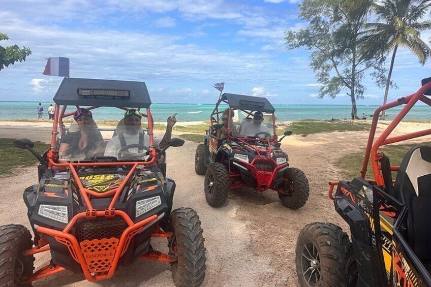 Three off-road vehicles parked on a sandy path near a beach with palm trees and the ocean in the background. 2