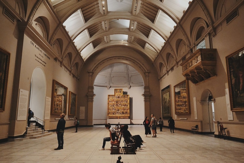 Une galerie d’art aux hauts plafonds et aux fenêtres en arc de cercle, où l’on peut admirer une grande peinture centrale ainsi que plusieurs œuvres d’art de plus petite taille accrochées aux murs. 1