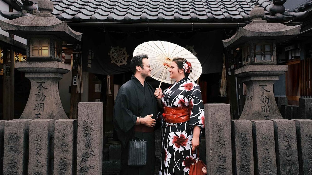 A couple in traditional Japanese attire standing in front of a temple with stone pillars and a tiled roof. 2
