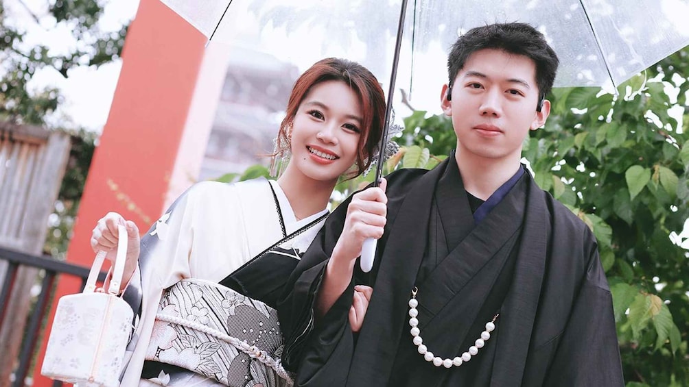 Two people in traditional attire, one holding a patterned bag and the other a pearl necklace, standing outdoors with greenery and a building in the background. 4