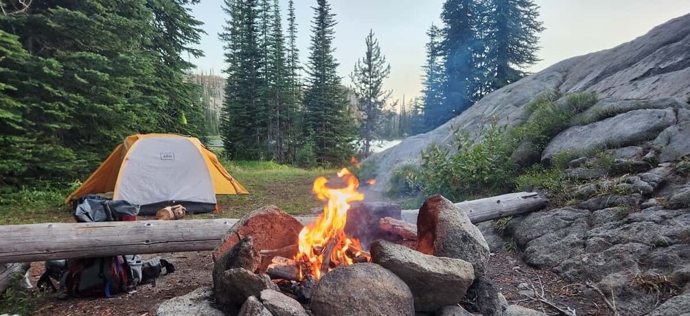 A campfire with a tent and gear nearby in a forested area. 1