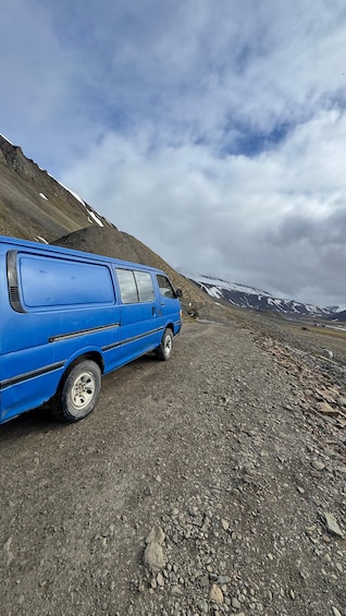 A blue van parked on a rocky mountain road with snow-capped peaks in the background. 2