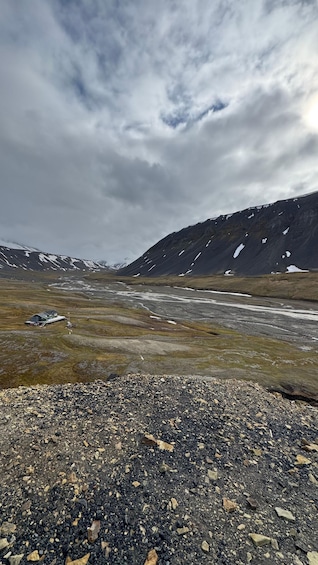 A gravelly foreground, a river, and snow-capped mountains under a cloudy sky. 5