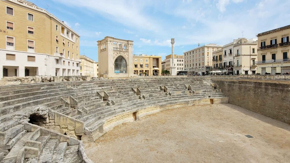 An ancient amphitheater with stone steps and a central stage, surrounded by modern buildings. 4