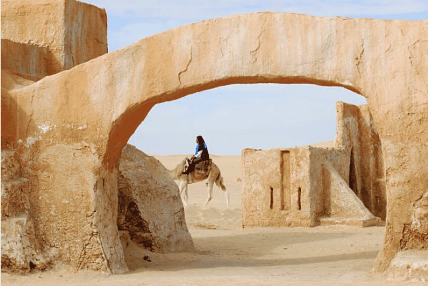 A person riding a camel under a large stone archway in a desert landscape. 3
