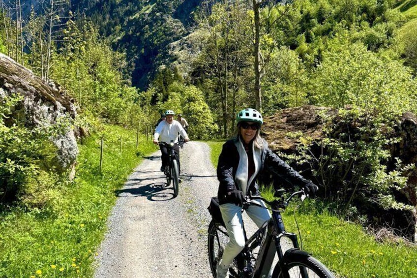 Two cyclists on a narrow mountain road surrounded by lush greenery and rocky terrain. 2