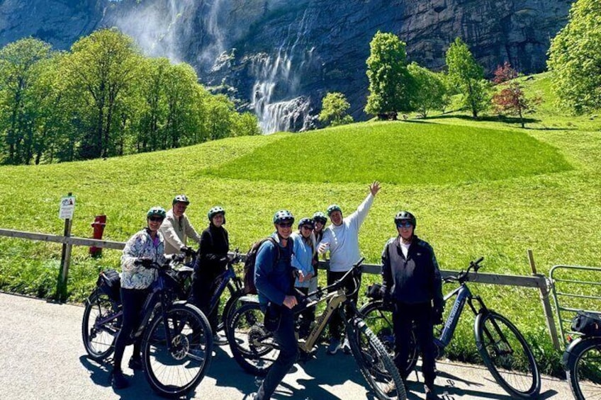 A group of cyclists on a paved path with a waterfall and green hills in the background. 1
