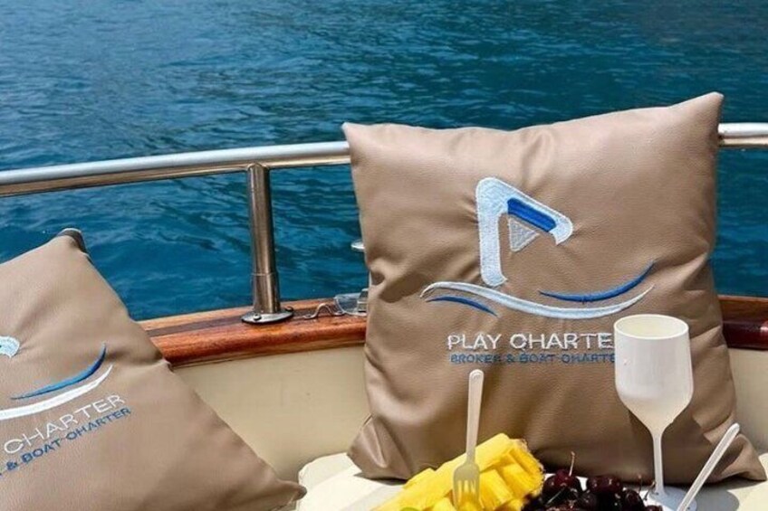 Two pillows with 'PLAY CHARTER BROKER & BOAT CHARTER' text, a glass of water, and a yellow napkin on a boat. 4