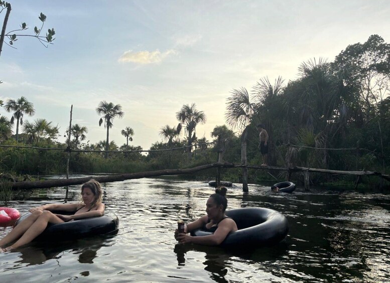 Two people floating on an inflatable tube in a river surrounded by palm trees and a wooden bridge. 2