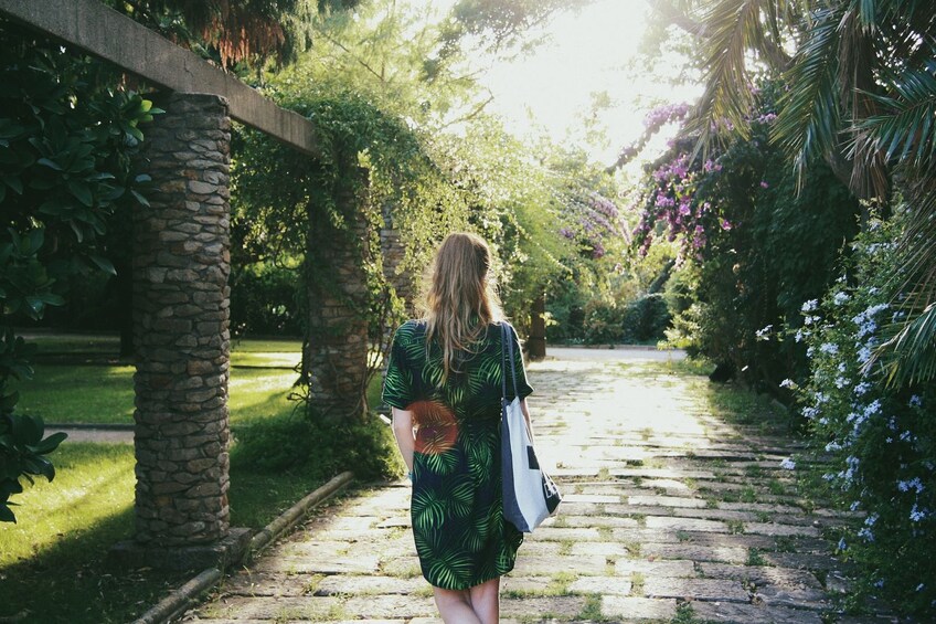 A woman walking down a stone pathway in a garden with lush greenery and flowering plants. 5