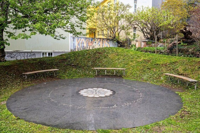 A circular paved area with benches, surrounded by grass and trees, with a building and graffiti in the background. 4
