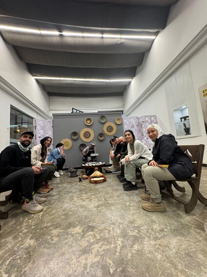 A group of people in a room with a grey ceiling, a table with woven baskets, and a display of kitchen utensils on the wall. 3