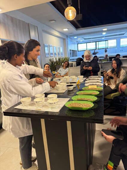 A group of people in a modern office setting, some standing and some seated, engaged in a discussion around a table with labeled cups and bowls. 2