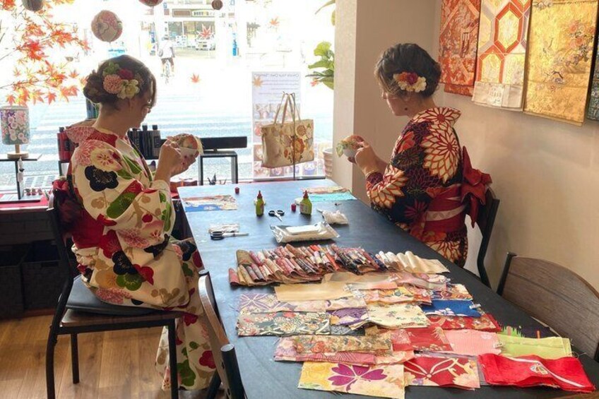 Two people in traditional attire at a table with various fabrics and a bottle. 5
