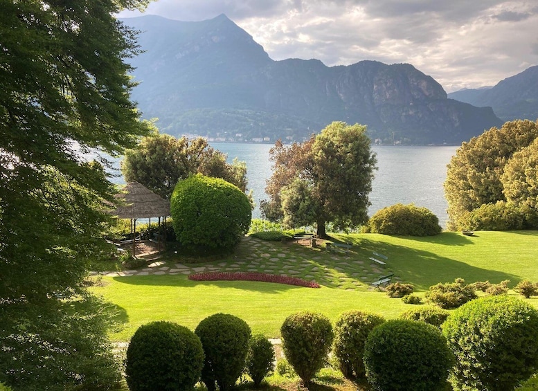 A landscaped garden with manicured bushes, a thatched gazebo, and a view of a lake with mountains in the background. 5