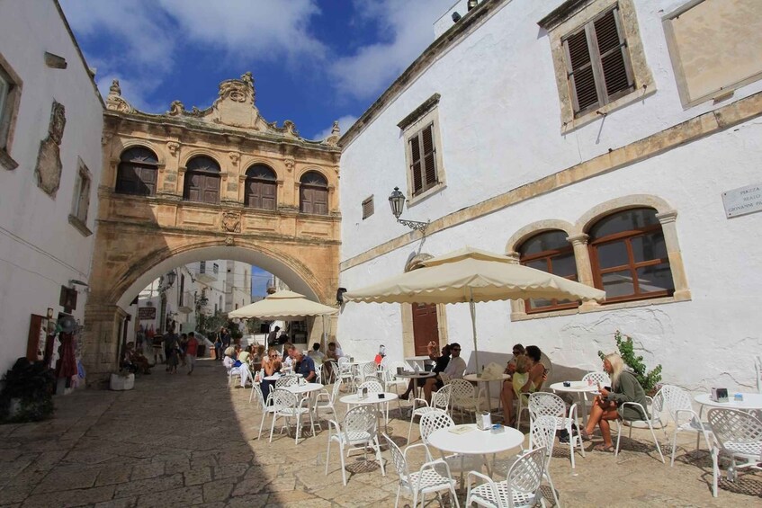 A cobblestone street with white buildings, outdoor seating, and an ornate archway. 1