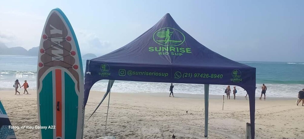 A beach with a Sunrise Rio Sup tent, a surfboard, and people walking along the shore. 4