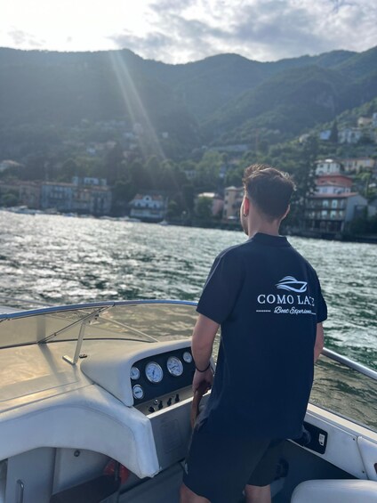 A person on a boat with 'COMO LAE Boat Experiences' on the back of their shirt, looking at a coastal town with buildings and hills in the background. 3