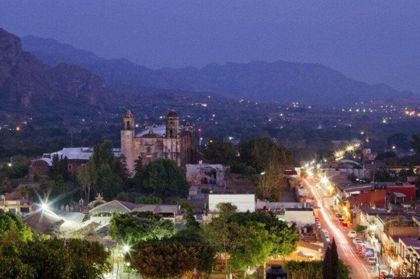 A cityscape at dusk with a prominent church, a mountain range in the background, and a busy street with vehicles. 4