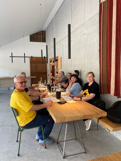 A group of people seated at a wooden table outdoors, with a concrete wall and a wooden shelf in the background. 5