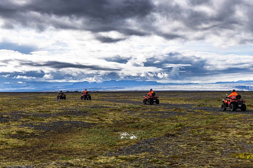 Four people on all-terrain vehicles in a barren landscape with distant mountains and a cloudy sky. 1