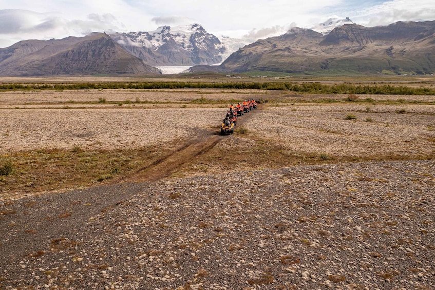 A convoy of orange vehicles on a gravel road with mountains and a glacier in the background. 4