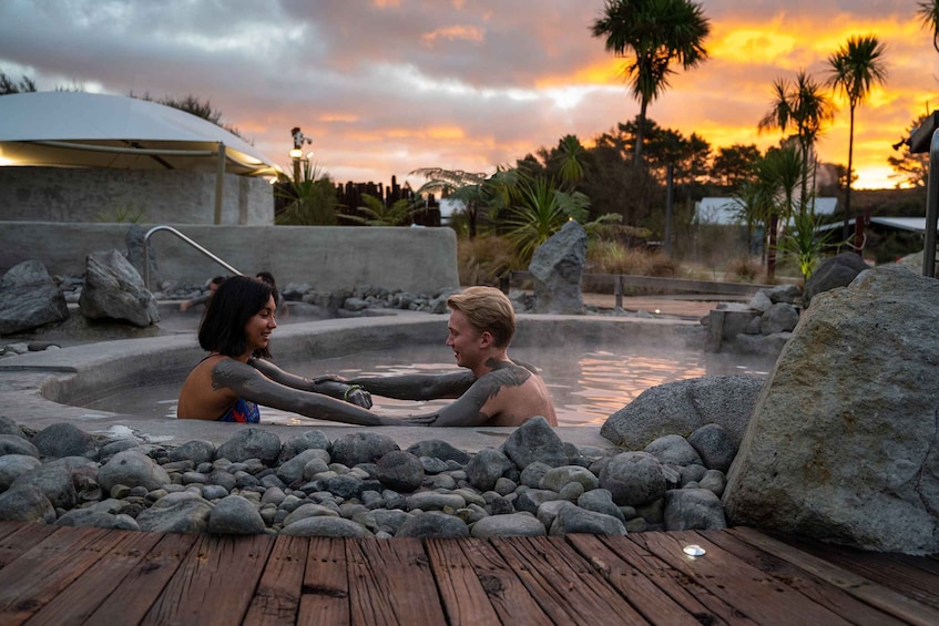 A couple in a hot spring surrounded by rocks and palm trees during sunset. 1